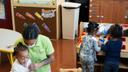 Children and a staffer in a daycare center. The children are gathered around desks and play areas.